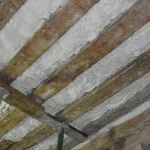 Imagen detallada del techo de un edificio antiguo, mostrando vigas de madera con aislamiento de espuma aplicado entre ellas, evidenciando una etapa intermedia de la restauración.