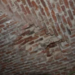 Vista cercana de una bóveda de ladrillo con signos de deterioro y daño estructural. Se nota la textura y el patrón de los ladrillos, con algunos agrietados y fragmentos faltantes, lo que indica la necesidad de reparación.
