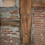 Imagen que muestra un detalle de la intersección entre una pared de ladrillo y un pilar de madera dentro de un edificio antiguo. La madera, con signos de desgaste y edad, contrasta con la mampostería deteriorada que la rodea.